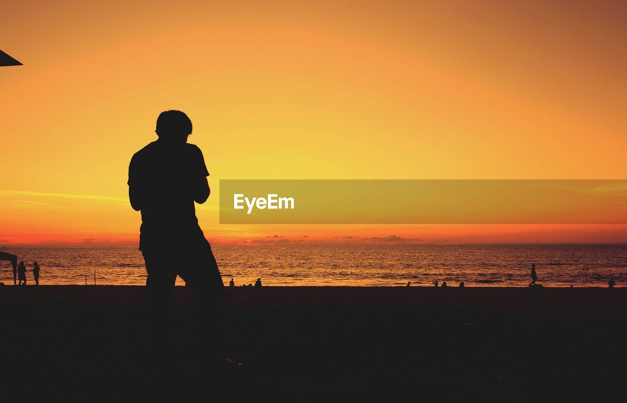 Silhouette man standing at beach against orange sky