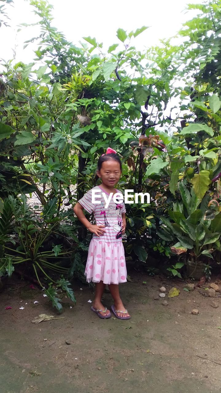 FULL LENGTH PORTRAIT OF GIRL STANDING AGAINST PINK AND PLANTS