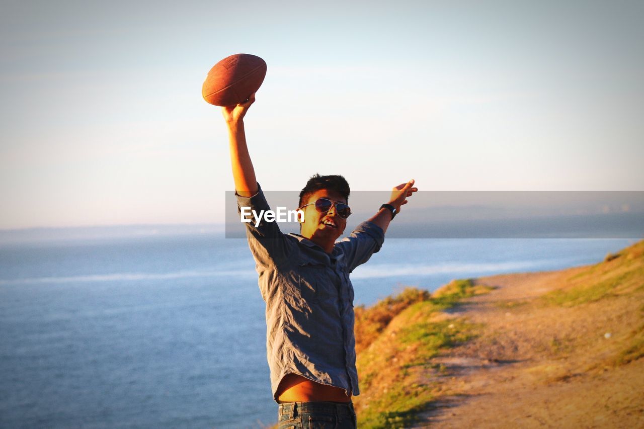 Young man with arms outstretched holding ball against sea