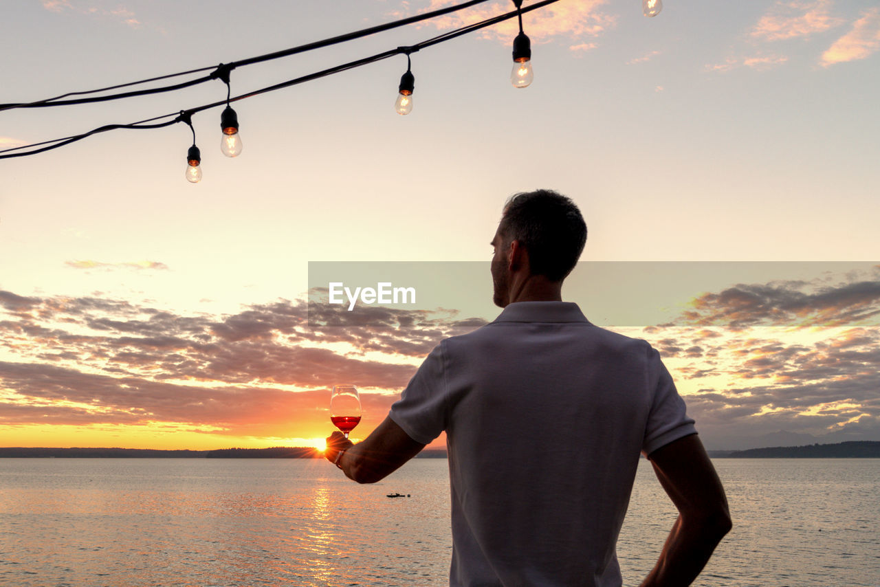 Rear view of mature man having wine against sea during sunset