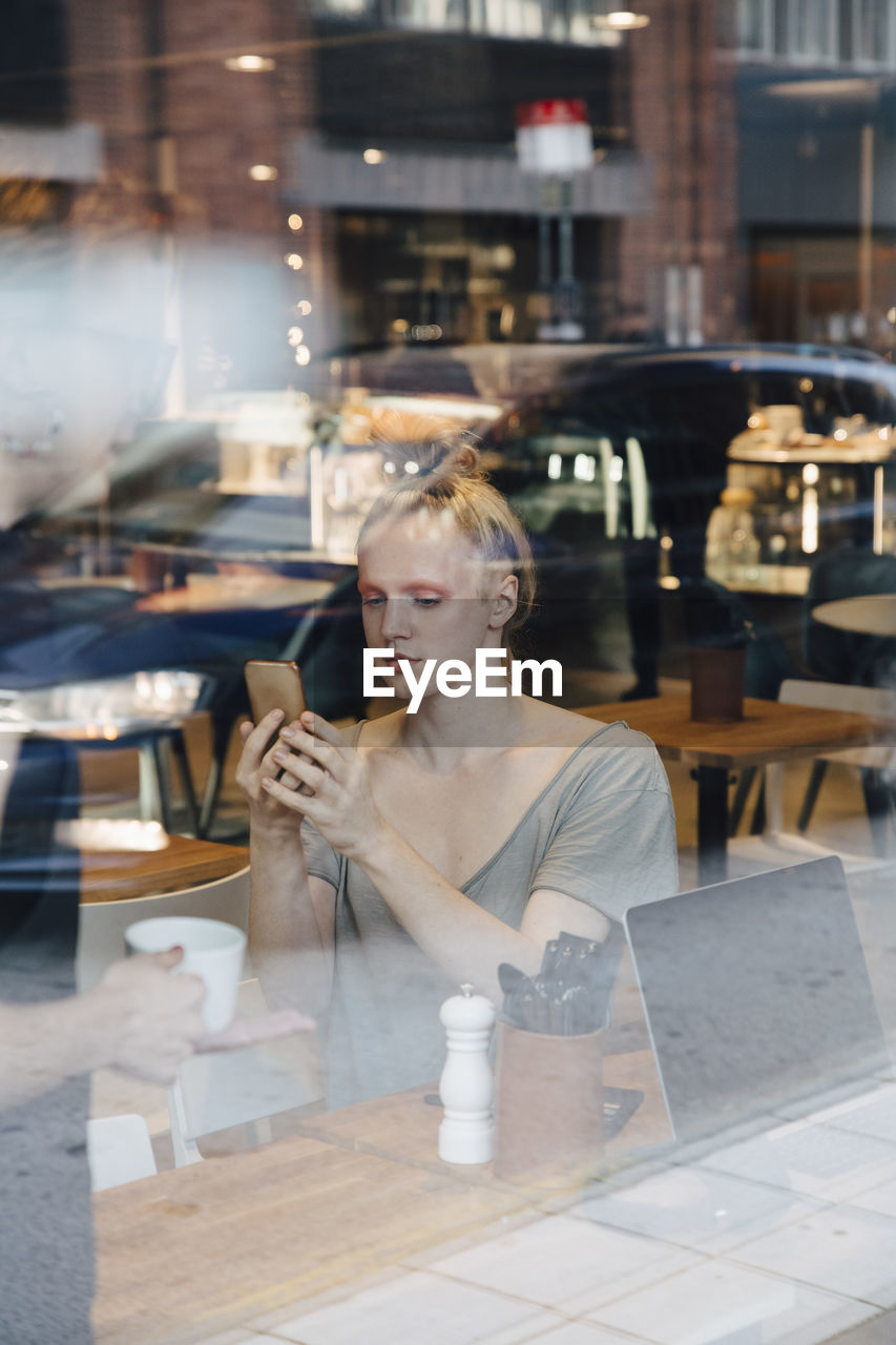 Young androgynous customer using smart phone while sitting in cafe seen from window