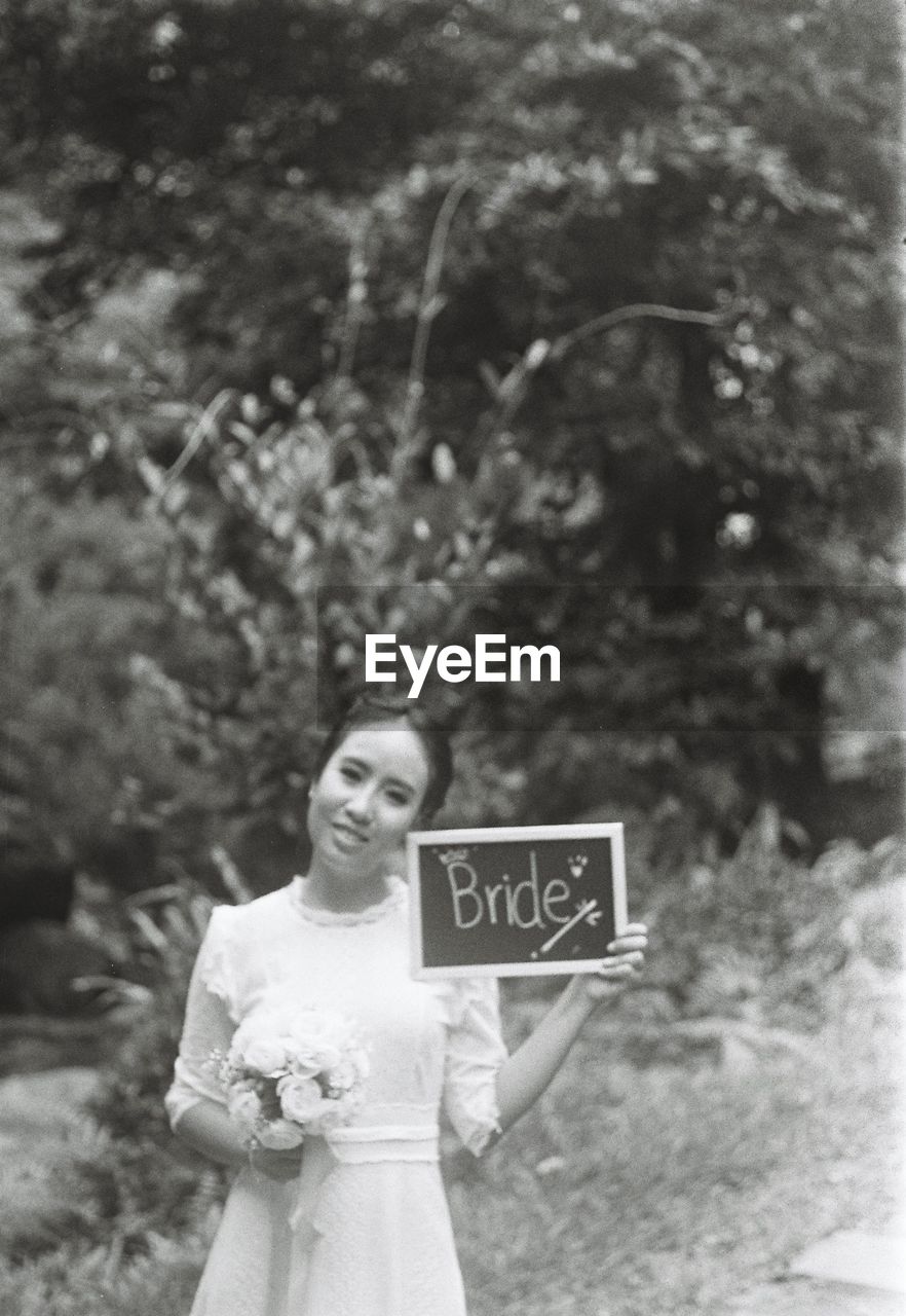 Portrait of bride holding slate with text and flower bouquet while standing against trees