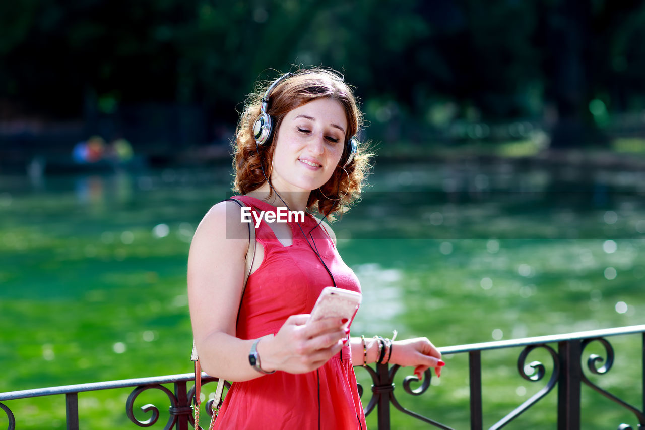 Smiling young woman using phone while standing against railing