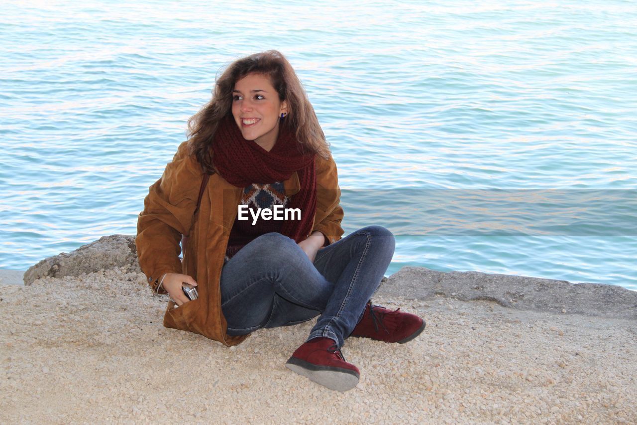 PORTRAIT OF SMILING WOMAN SITTING ON SHORE