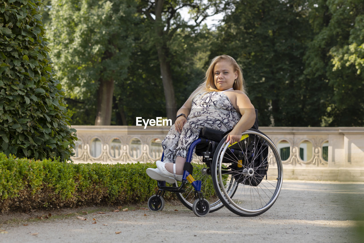 Full length young pretty woman with short stature on wheelchair looking at camera in public park at