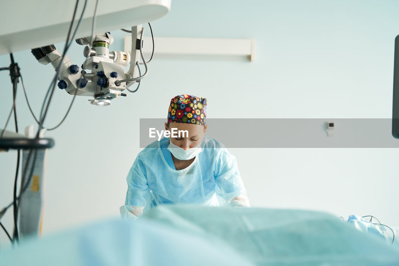 Attentive adult female medic in disposable mask and ornamental cap working in clinic with surgical microscope