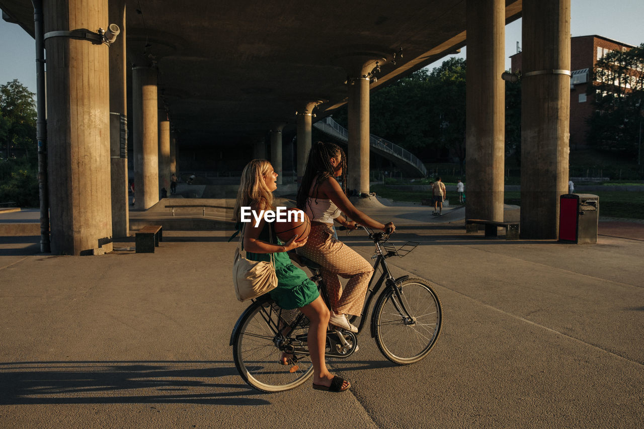 Side view of teenage girl sitting with friend riding bicycle on road during sunny day