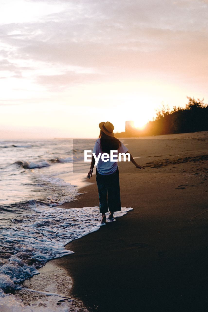 Rear view of woman standing at beach during sunset