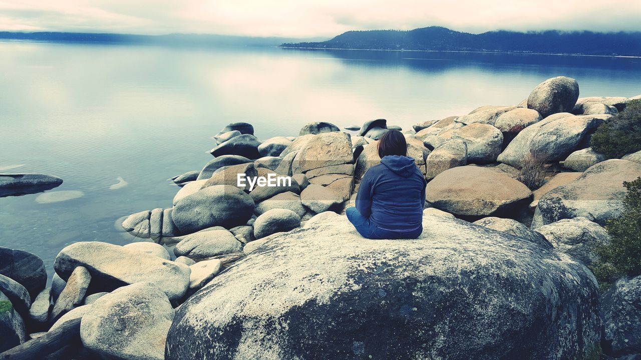 Rear view of person sitting on rock by river