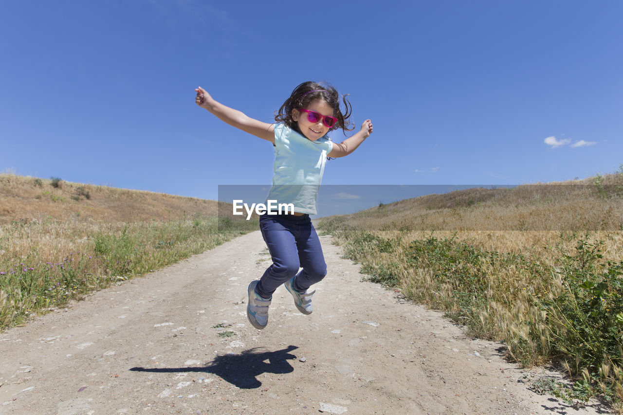 Full length of cheerful girl jumping on road amidst grassy field against sky