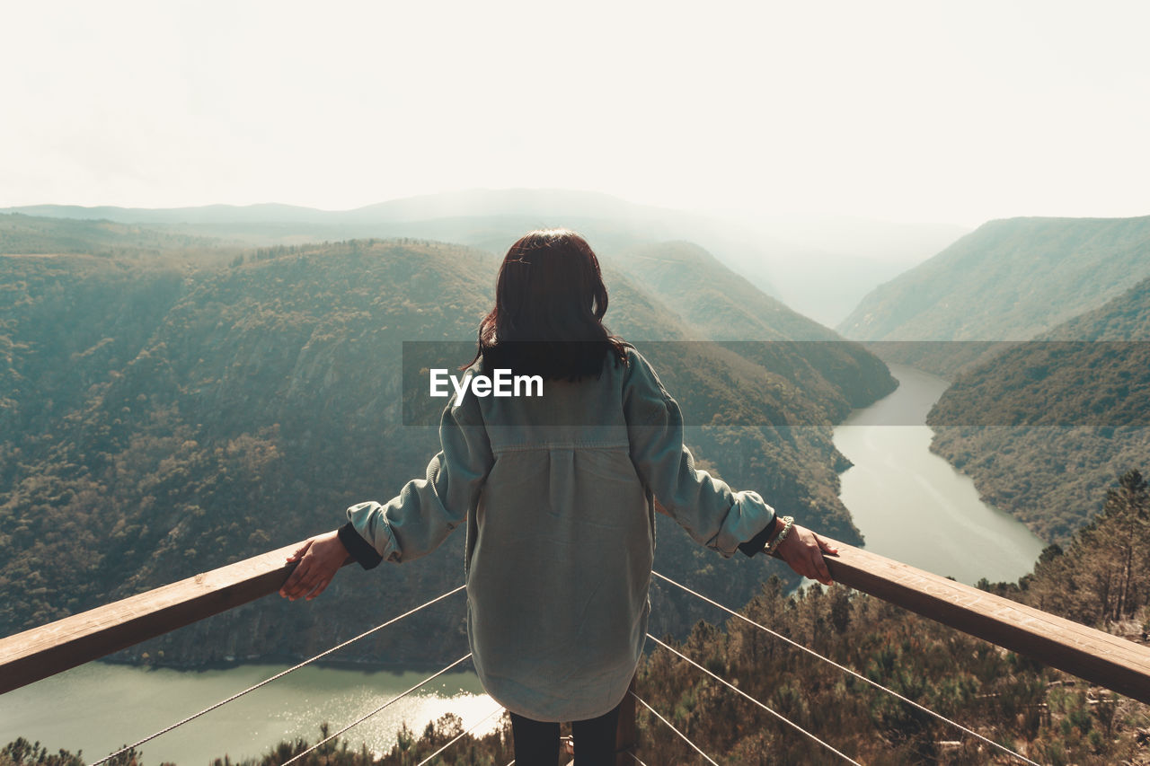 REAR VIEW OF WOMAN STANDING ON RAILING AGAINST MOUNTAIN