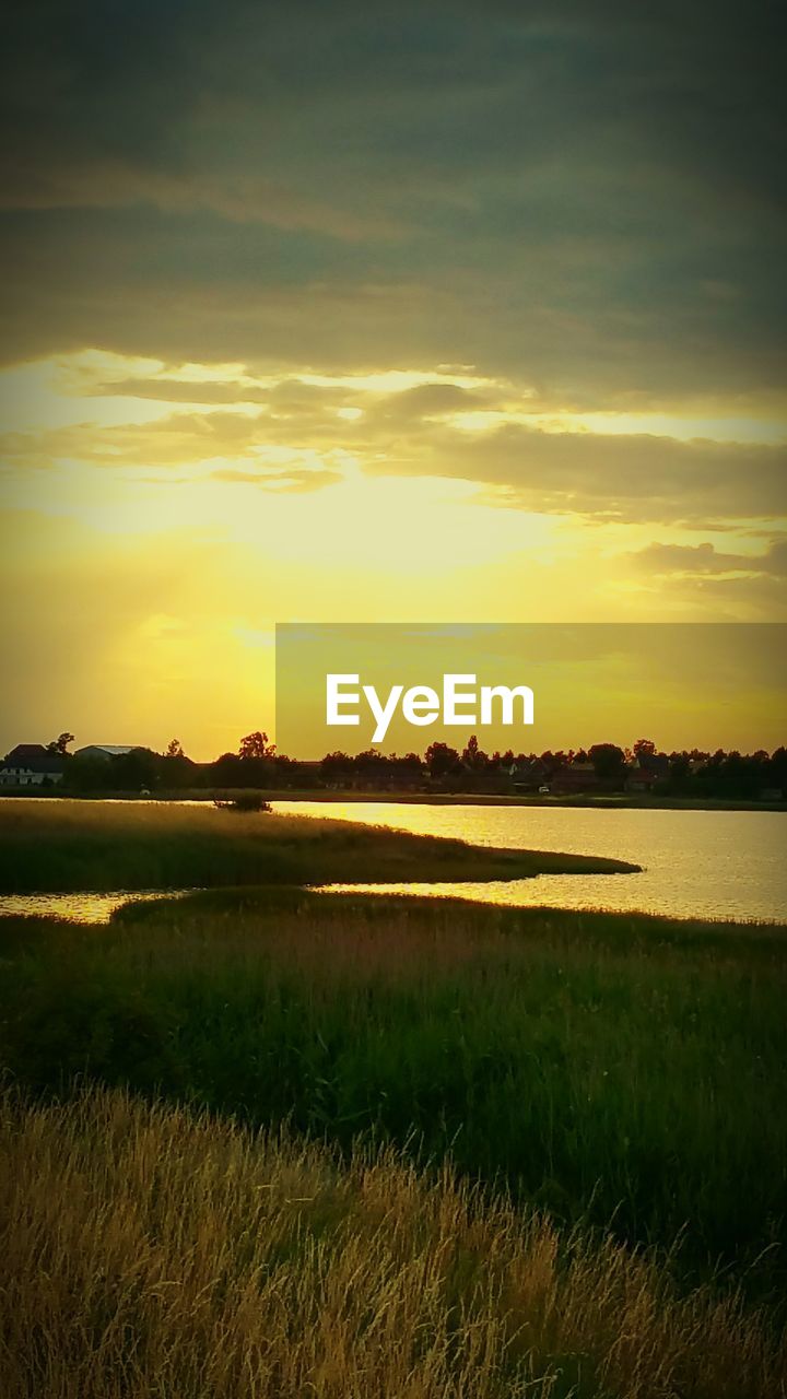 Scenic view of grass field by river against cloudy sky during sunset