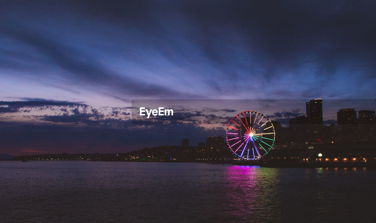 Illuminated ferris wheel by sea against sky at night