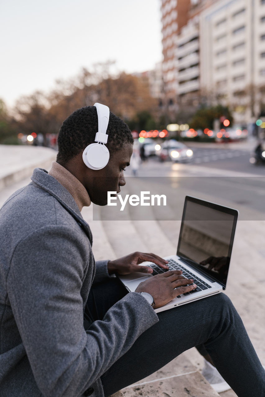 Young man wearing headphones working on laptop while sitting in city