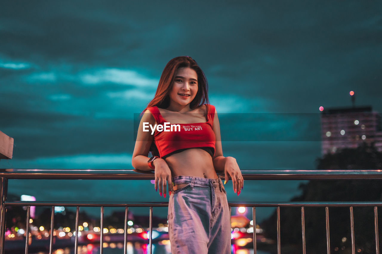 Low angle view of smiling young woman standing on bridge against sky at night