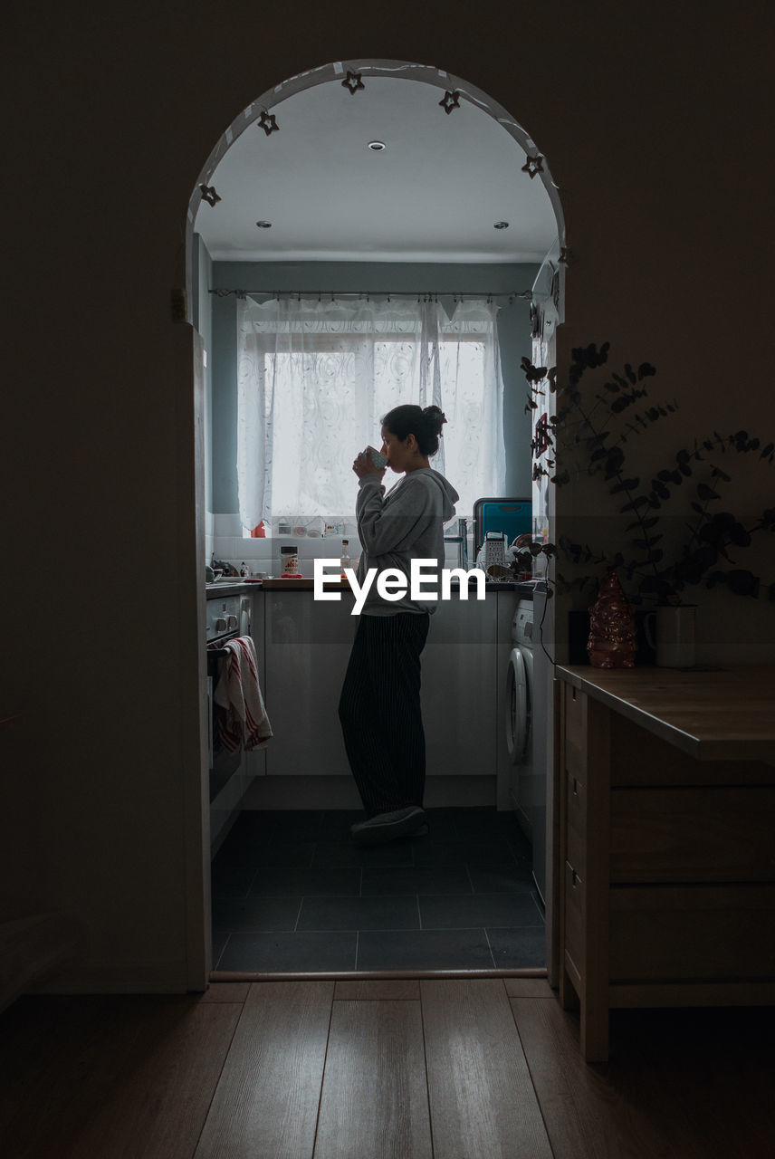 REAR VIEW OF WOMAN STANDING ON WINDOW AT HOME