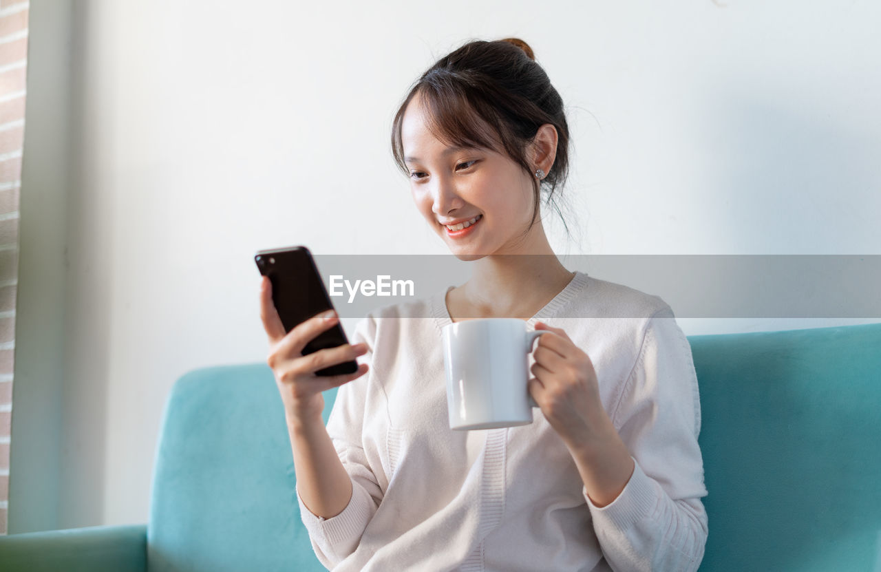 young woman using mobile phone while standing against wall