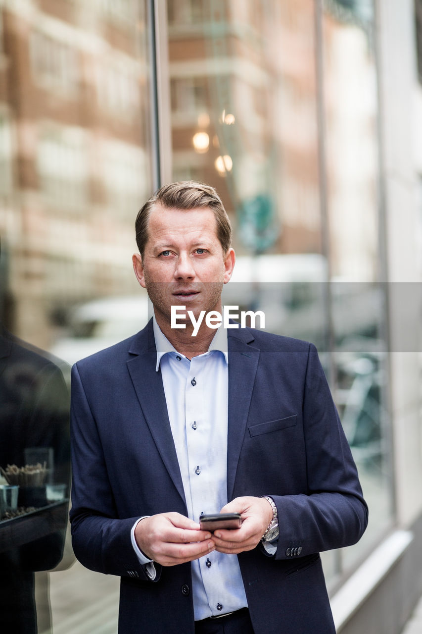 Contemplating mature businessman with smart phone looking away while standing by glass window