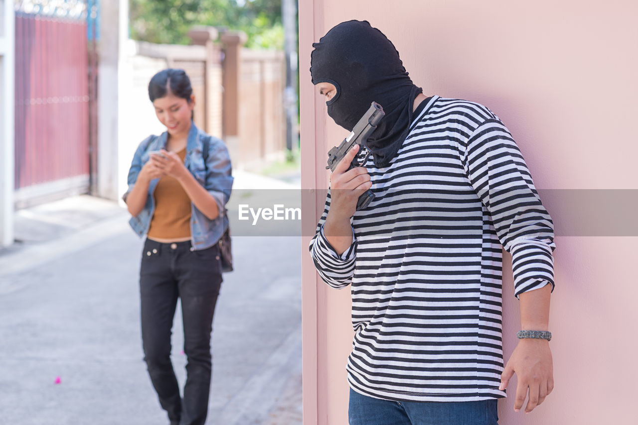 Thief with gun hiding against wall by smiling young woman walking on street by