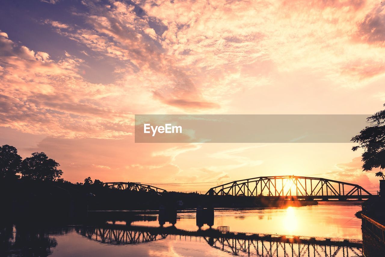 Silhouette bridge on river against cloudy sky during sunset