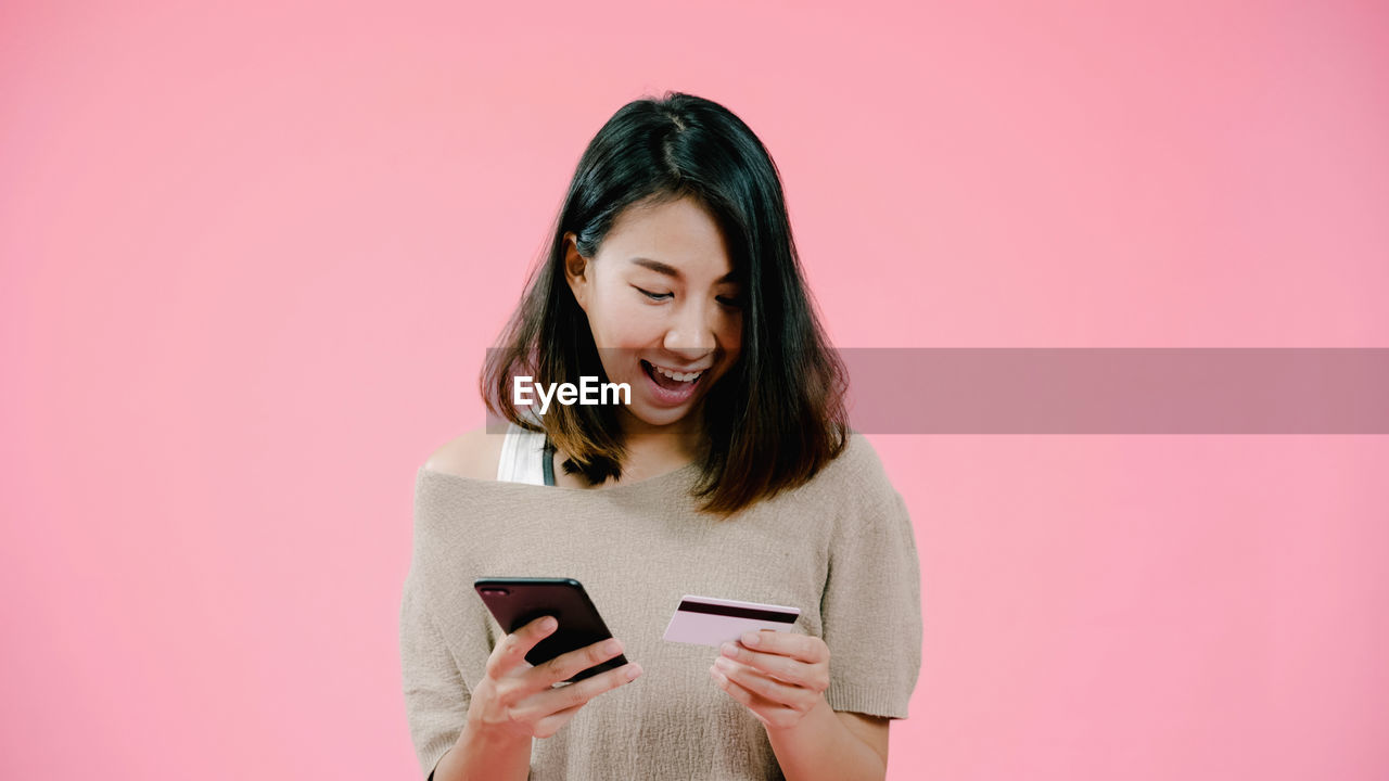 Smiling woman doing online shopping through mobile phone against pink background