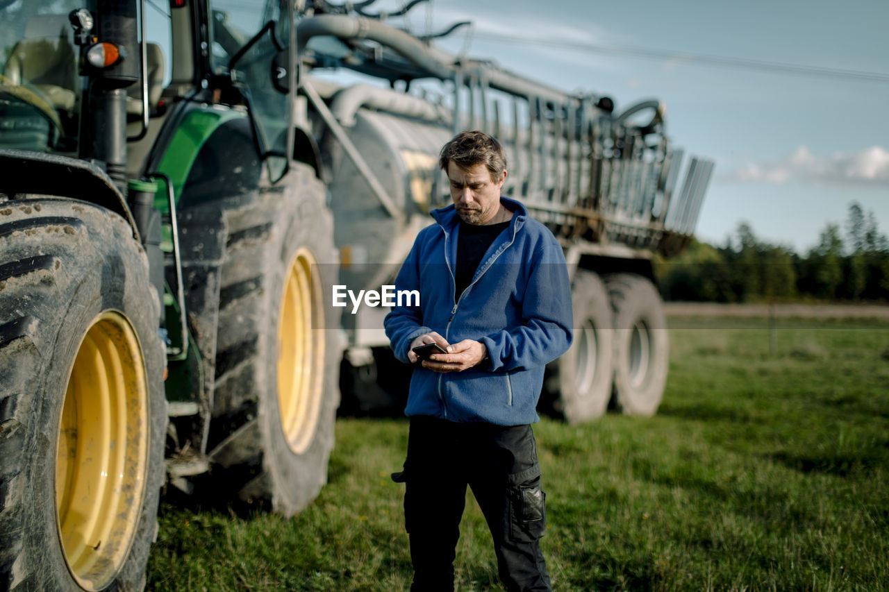 Mature farmer using smart phone standing against tractor on field