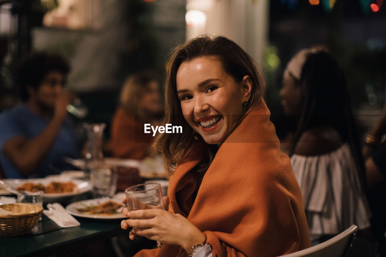 Portrait of smiling young woman holding drink while sitting at table during dinner party in backyard