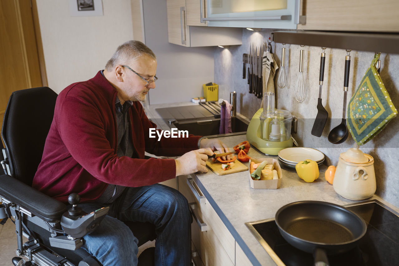 Retired senior man with disability sitting in motorized wheelchair cutting bell pepper at kitchen counter