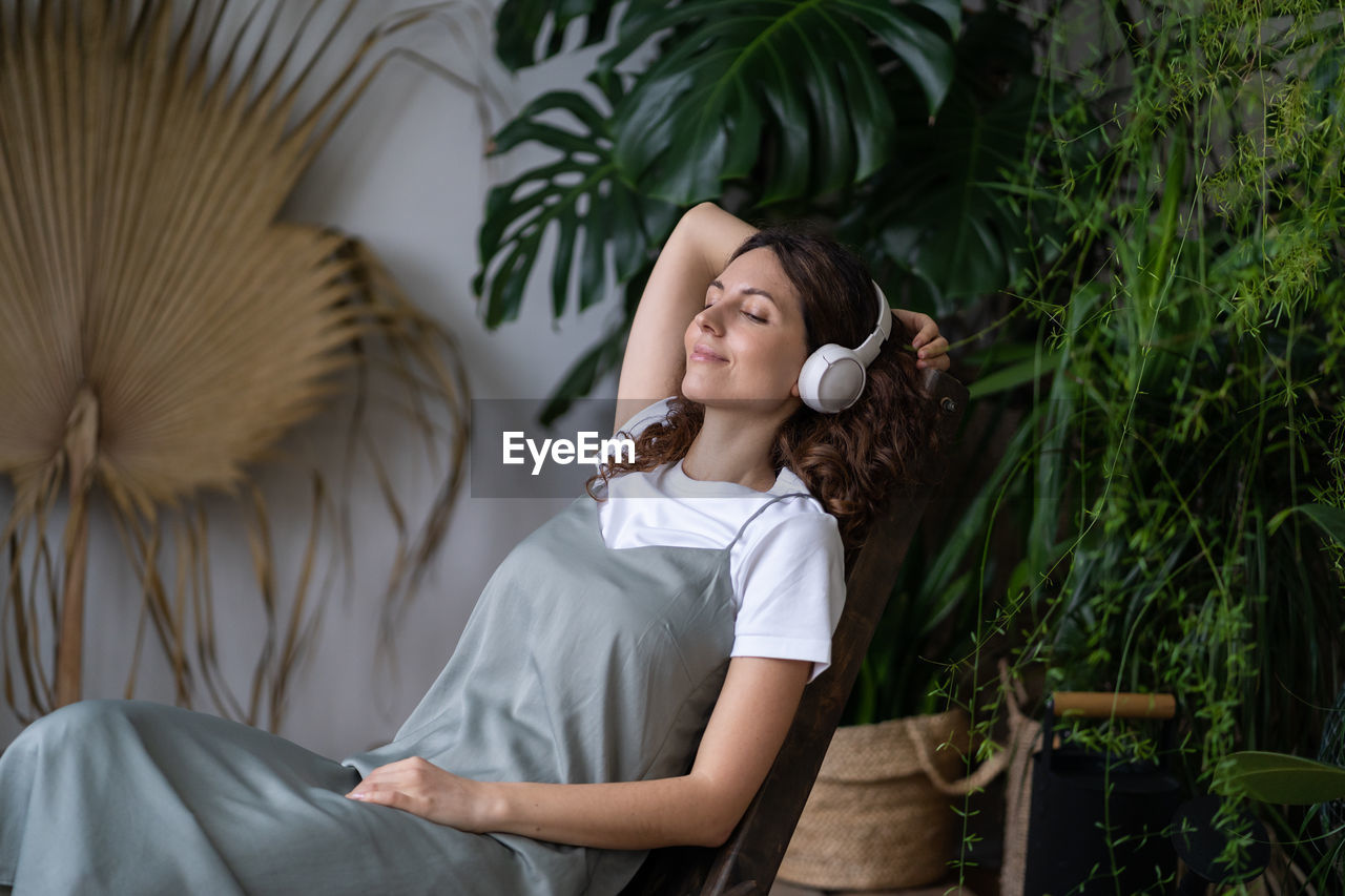 Calm young woman florist relax in house garden wearing wireless headphone after work with houseplant