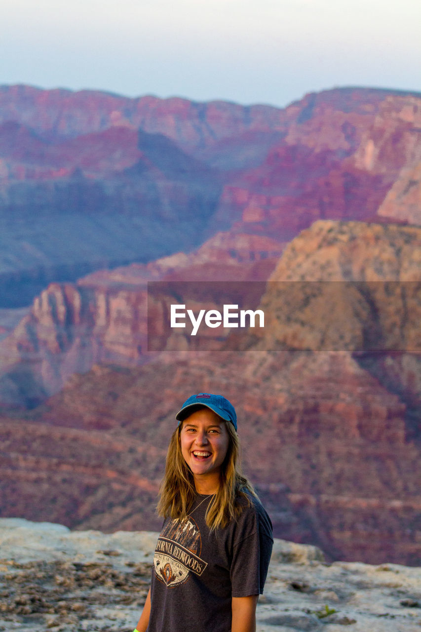 PORTRAIT OF SMILING WOMAN STANDING AGAINST MOUNTAINS