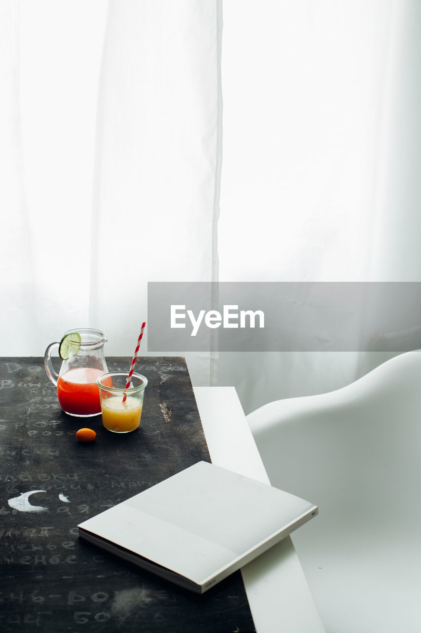 From above of jar and glass with red and orange fruit drinks placed with book on table near window in light room