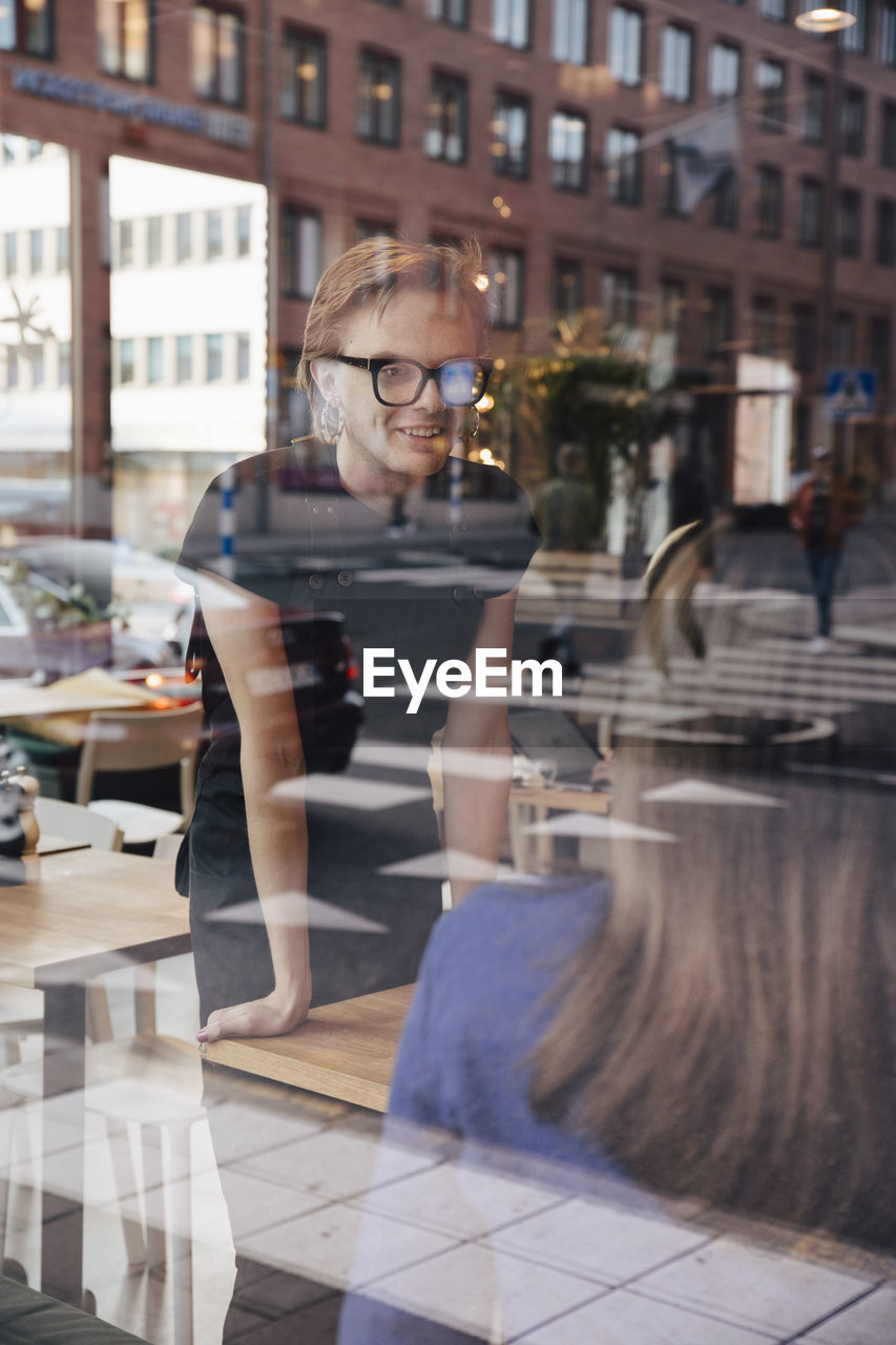 Smiling young transgender barista talking with female customer seen through cafe window