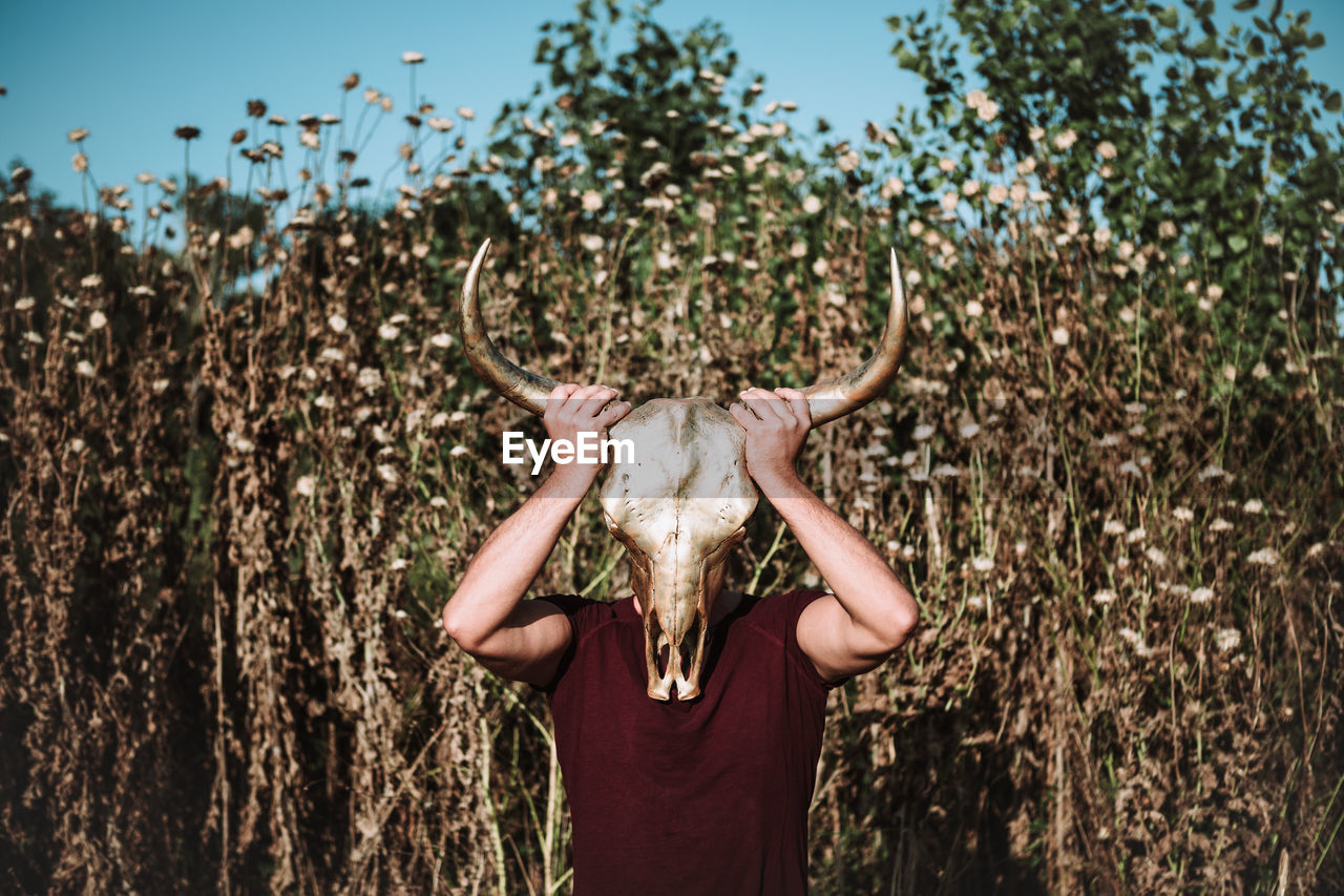 Full length of anonymous male in casual wear covering face with horn animal skull while standing in field