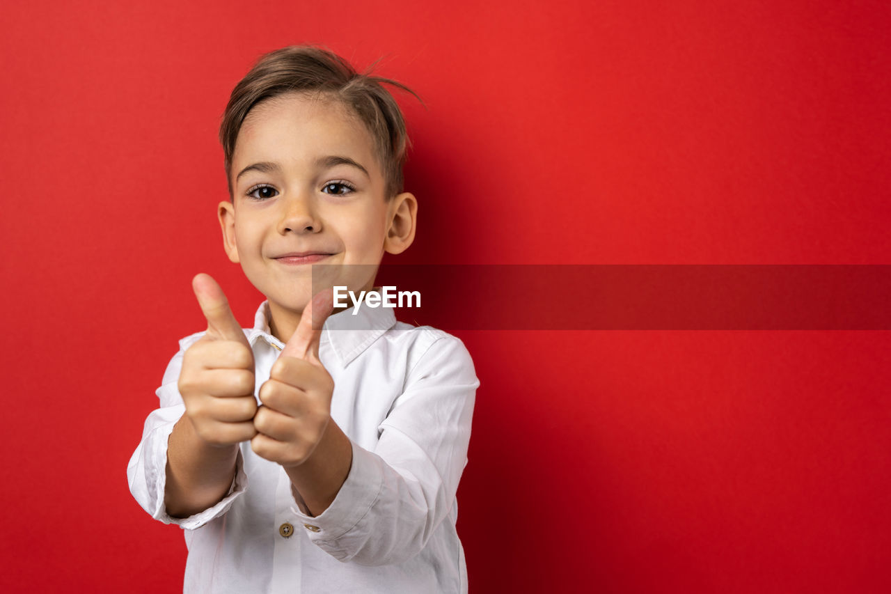 childhood, child, portrait, colored background, one person, studio shot, red, looking at camera, finger, smiling, hand, person, happiness, indoors, emotion, gesturing, baby, thumbs up, men, cute, red background, copy space, waist up, sign language, positive emotion, front view, showing, cheerful, human face, hand sign, toddler, fun, cut out, arm, human mouth, female, education, clothing, nose