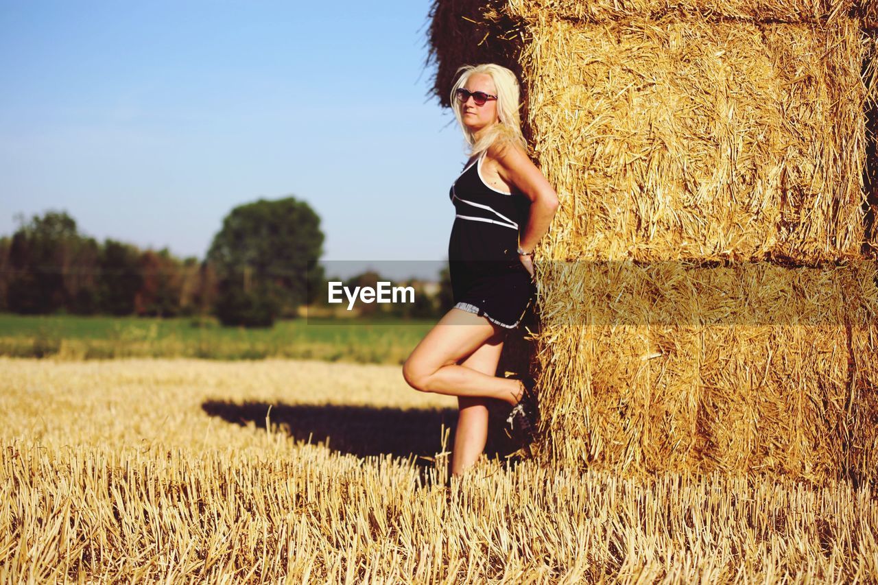 Side view of young woman wearing sunglasses standing by hay bales at farm