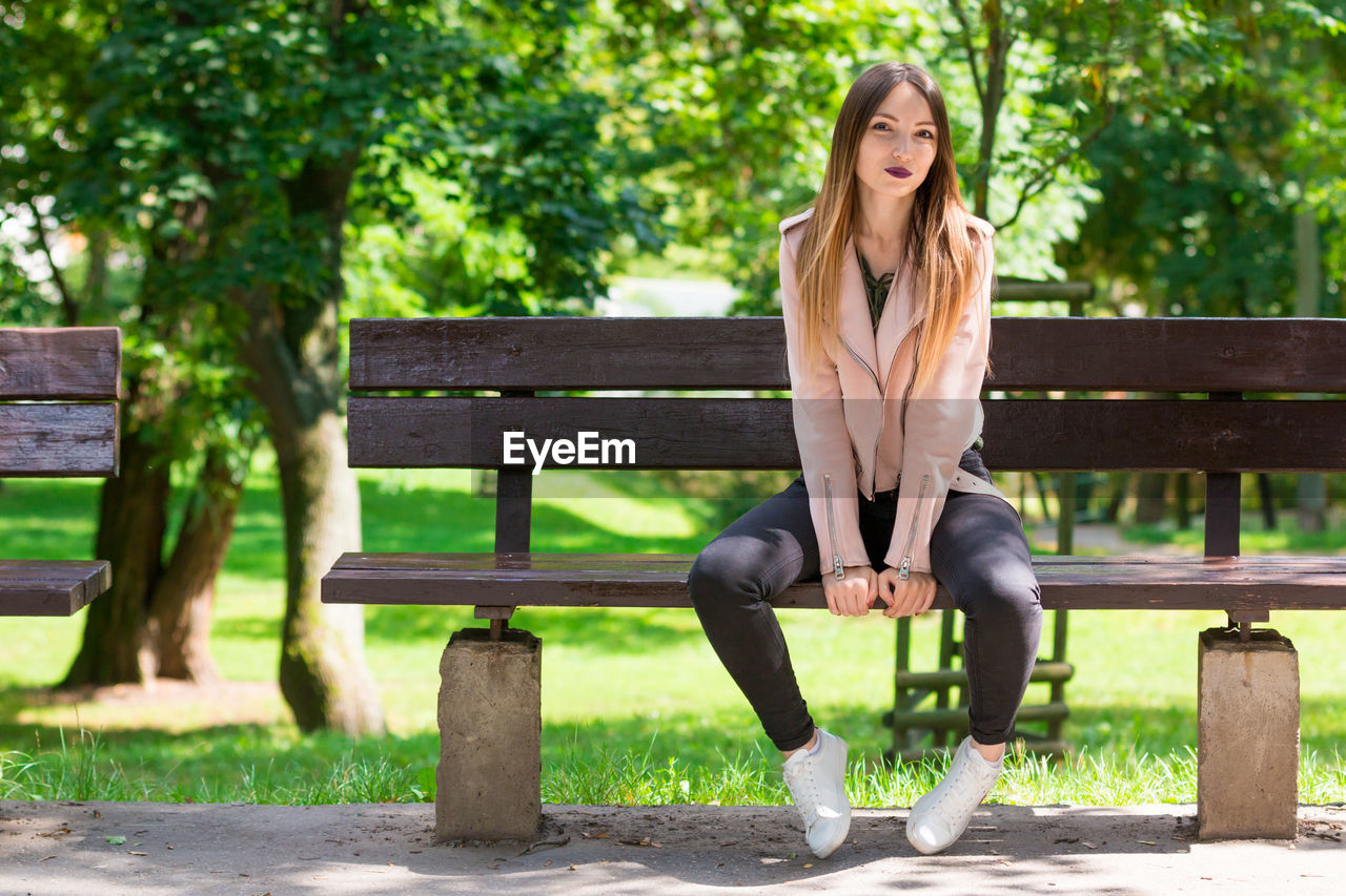 Full length portrait of beautiful woman relaxing on bench at park during sunny day