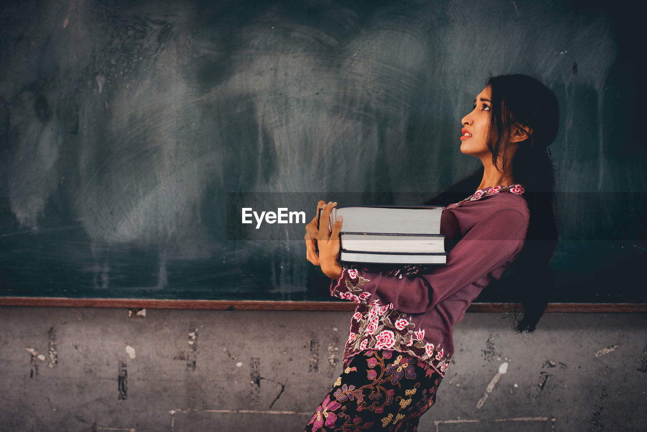 Side view of young woman with books looking up while standing by blackboard in classroom