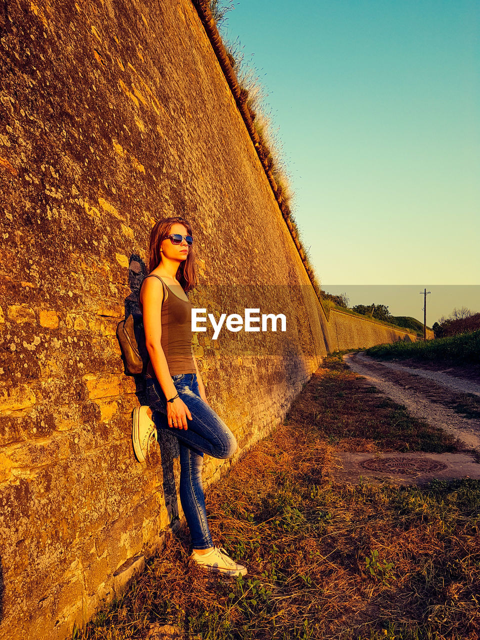 Side view of young woman standing on field by retaining wall against clear sky during sunset