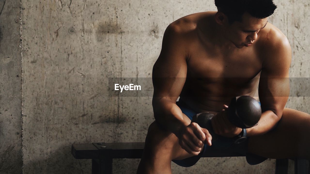 Shirtless man holding dumbbell while sitting in gym