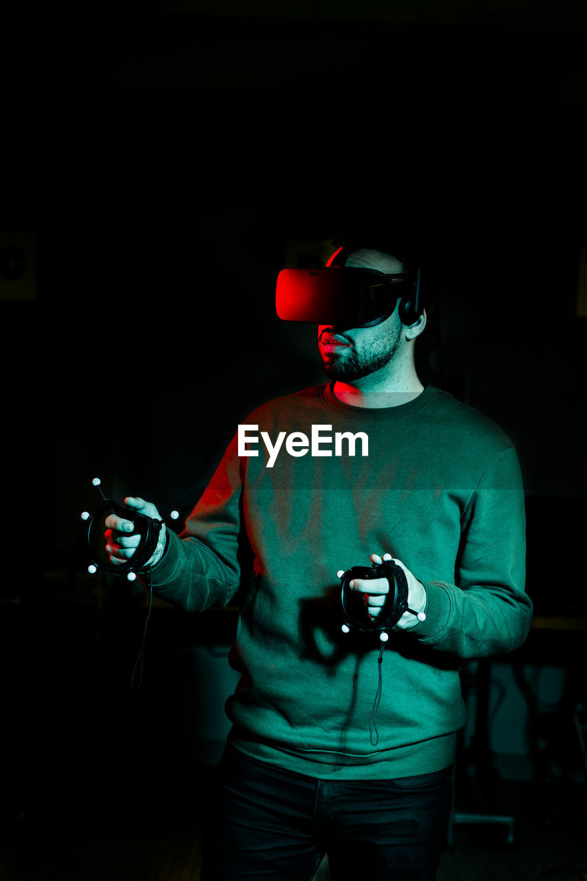 A young man wearing virtual reality glasses stands in a darkened room