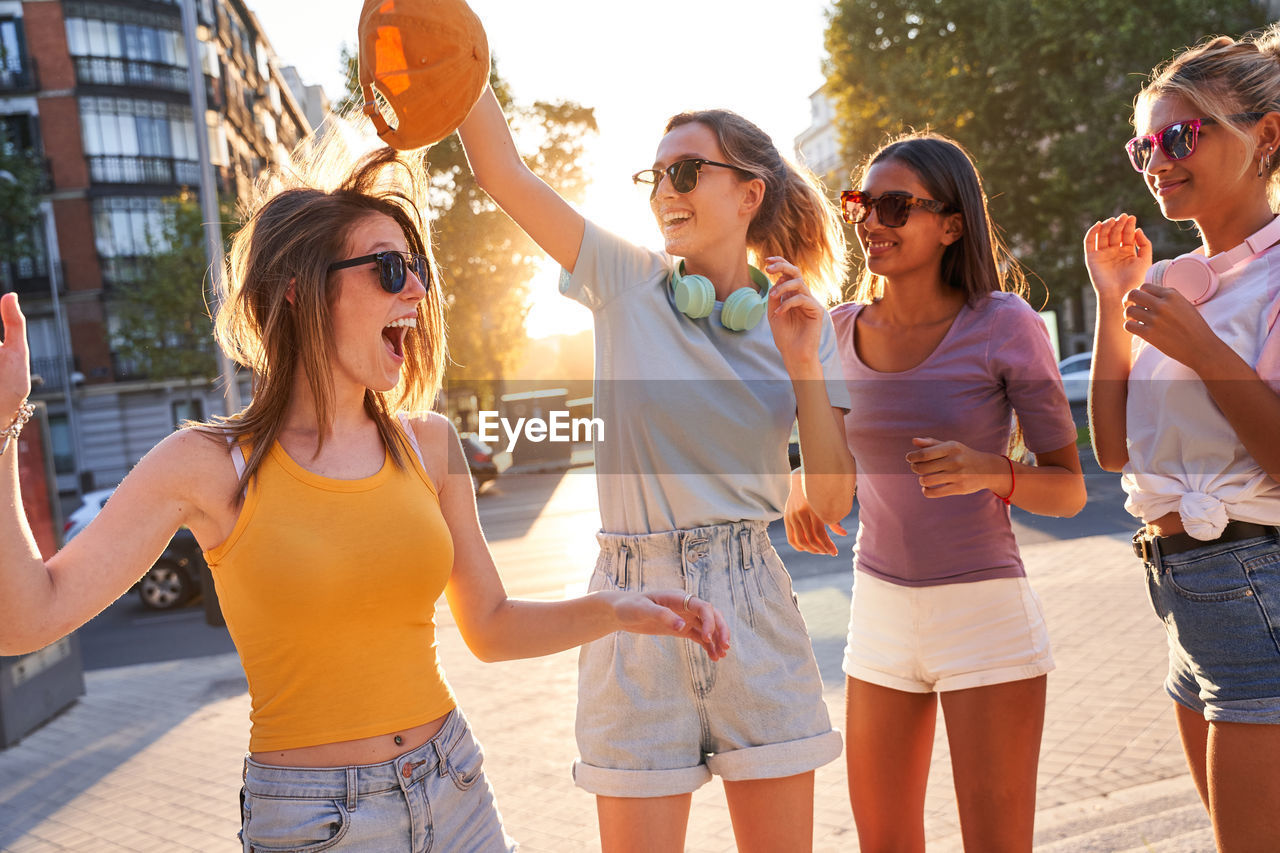 Happy carefree teen female friends in trendy clothes and sunglasses having fun and dancing together on urban street while enjoying summer day in city