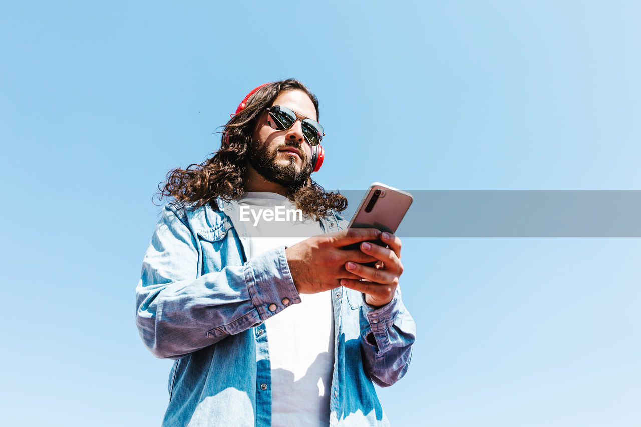 From below bearded latin american male in modern sunglasses browsing on cellphone and wireless headphones on blue background on sunny day