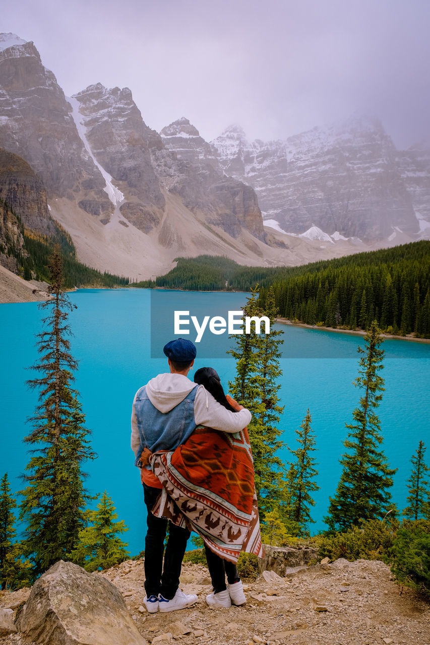 REAR VIEW OF MAN LOOKING AT LAKE AGAINST MOUNTAIN RANGE