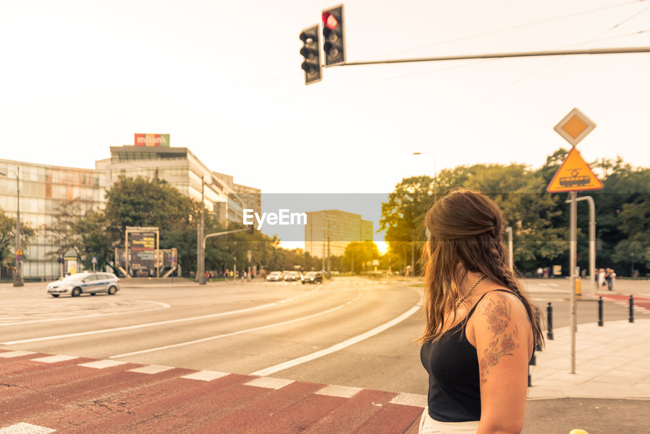 Young woman standing on city street against clear sky during sunset