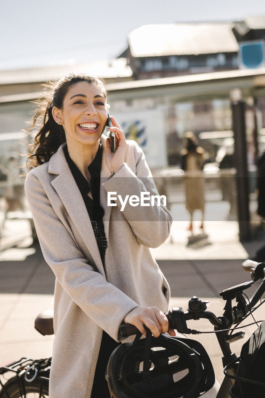 Happy businesswoman wearing long coat holding bicycle while talking on smart phone