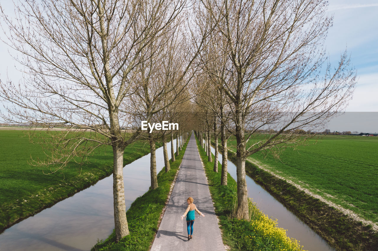 REAR VIEW OF MAN WALKING ON FOOTPATH AMIDST BARE TREES