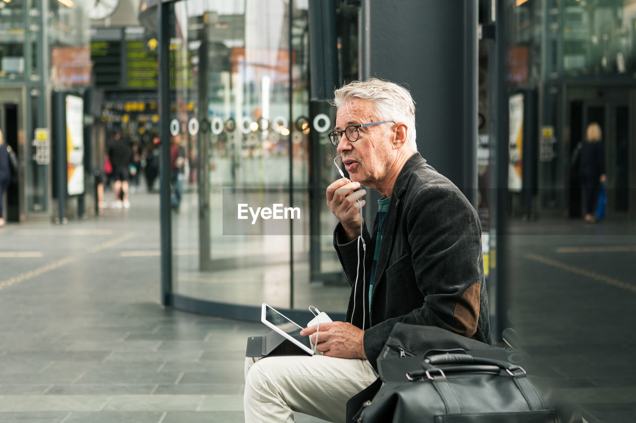 Senior male commuter talking through in-ear headphones while sitting by bags at railroad station