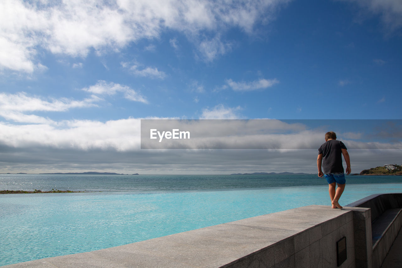 REAR VIEW OF MAN STANDING AGAINST SEA