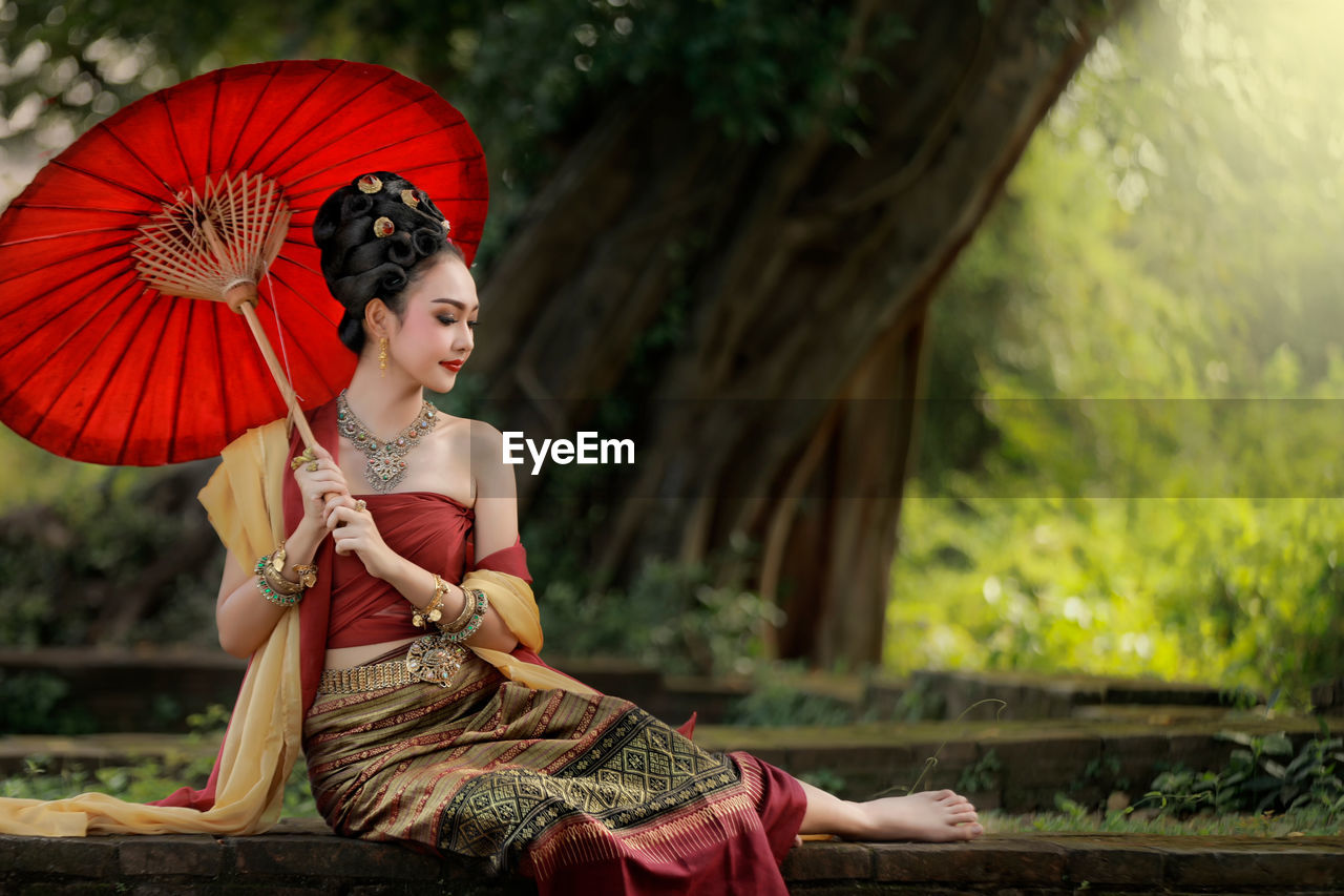 Young woman holding red umbrella while sitting on retaining wall