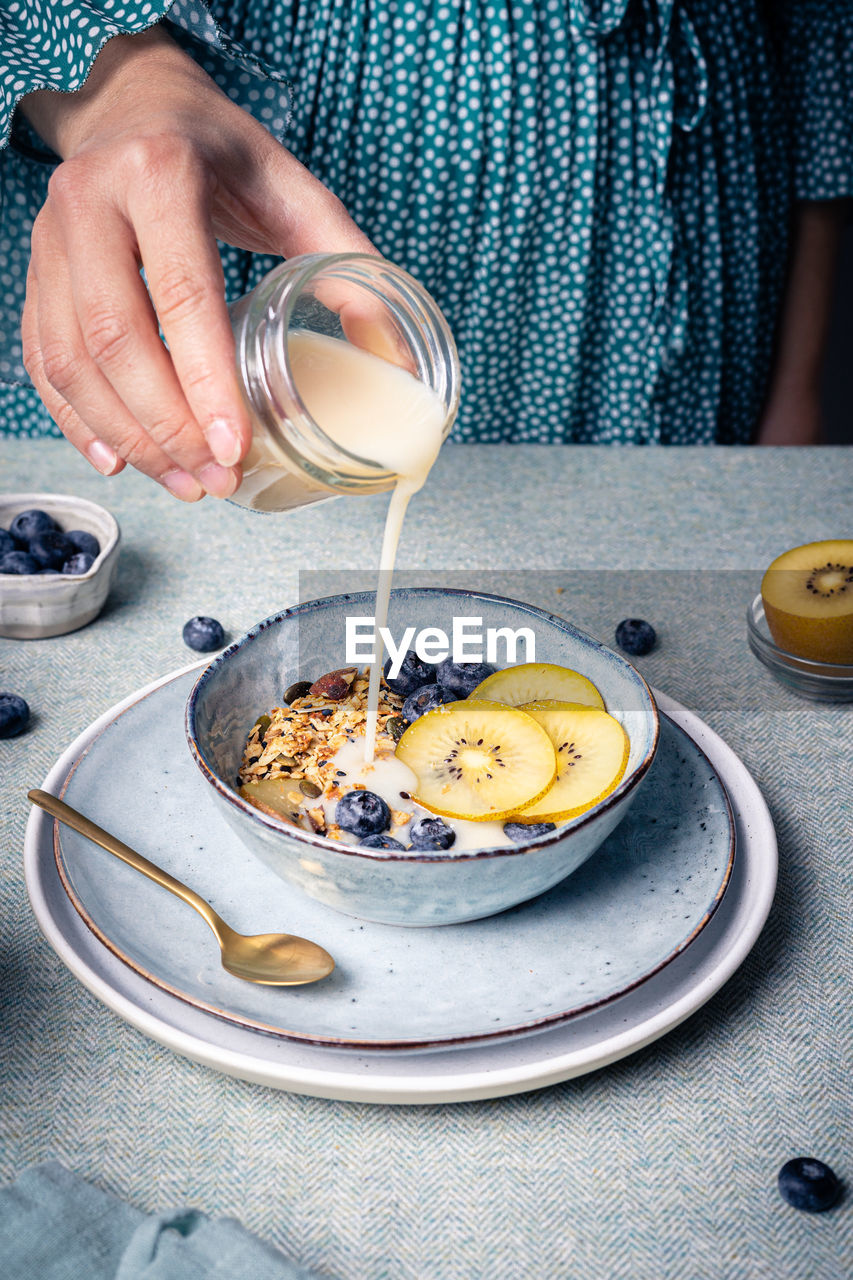 High angle of crop anonymous female adding milk into bowl with healthy muesli with blueberries and pieces of kiwi for healthy breakfast